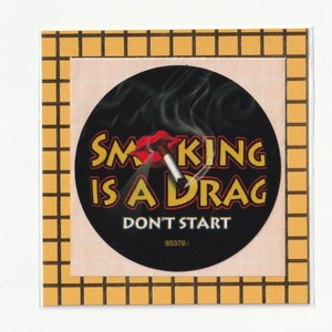 May include: A square sticker with a black circle featuring a dart piercing a cigarette, with smoke rising. The text reads "SMOKING IS A DRAG DON'T START" in yellow and white. The sticker has a yellow and black grid border.