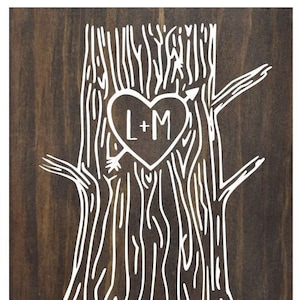 Pode incluir: Uma placa de madeira com um contorno branco de uma árvore com um coração esculpido no tronco. O coração tem as iniciais "L+M" dentro. O texto "Lacey & Mike" e "NOVEMBER 2, 2019" está abaixo da árvore.