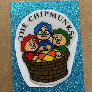 May include: A colorful sticker featuring the Chipmunks cartoon characters in a basket. The sticker is on a blue glitter background. The Chipmunks are in a brown basket with a checkered pattern.