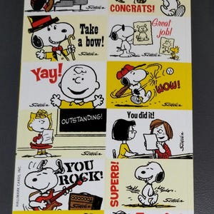 May include: A sheet of colorful stickers featuring Peanuts characters in various poses with phrases like "Here's to you!", "Congrats!", and "You Rock!" The stickers have a yellow background and are from Hallmark Cards.