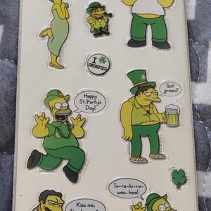 May include: Sheet of metallic stickers featuring The Simpsons characters in St. Patrick's Day attire. Homer, Marge, and other characters are depicted with green hats, beer, and speech bubbles with phrases like "Happy St. Party's Day!" and "Beer me, I'm Irish."