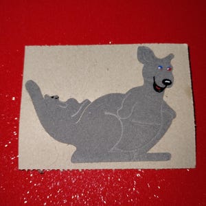 May include: A gray kangaroo sticker with blue and red eyes and a black mouth. The sticker is on a rectangular piece of cardboard with a rough edge. The background is a solid red color.
