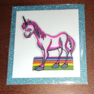 May include: A white unicorn with pink accents stands on a rainbow. The unicorn has a black horn and a gray mane. The rainbow is made of red, orange, yellow, green, blue, indigo, and violet.