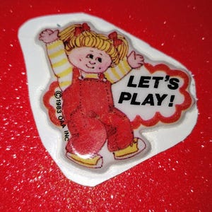 May include: A sticker featuring a cartoon girl with blonde hair wearing a red overall dress and a white shirt with yellow stripes. The sticker has a white background with a red cloud shape and the text "Let's Play!" in black letters. The copyright information "©1983 OAA INC" is printed in black letters at the bottom of the sticker.