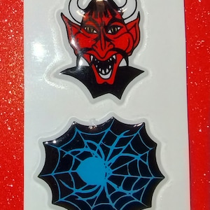 May include: Two Halloween-themed stickers. The top sticker features a red devil face with horns and a black collar. The bottom sticker shows a black spider web with a blue spider in the center. Both stickers are on a white background.
