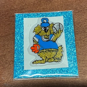 May include: A cartoon character wearing a blue baseball cap and jersey, holding a baseball and a glove. The character has brown fur and is set against a blue glitter background.