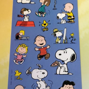 May include: A sheet of colorful stickers featuring various characters from the Peanuts comic strip. The stickers include Snoopy, Charlie Brown, Lucy, and Woodstock in various poses. The background is a light blue color.