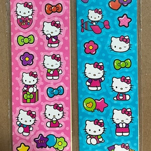 May include: Two sheets of Hello Kitty stickers. The sheet on the left is pink with white polka dots and features Hello Kitty in various poses with bows, hearts, stars, and a rainbow. The sheet on the right is blue with white polka dots and features Hello Kitty in various poses with bows, stars, hearts, and a plane. Both sheets have the text "Hello Kitty" on them.