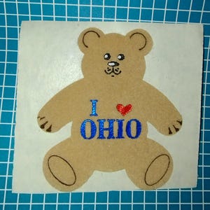 May include: A tan teddy bear sticker with the text "I ❤️ OHIO" in blue and red. The bear has black details for the eyes, nose, and claws. The sticker is on a white background.