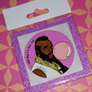 May include: A sticker featuring a cartoon image of Mr. T blowing a bubblegum bubble. The sticker is in a clear plastic package with a purple glitter border. The background is pink and orange.