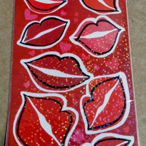 May include: A sheet of red lip stickers with a variety of red shades and black outlines. The stickers have a glossy finish and are arranged on a red background with heart-shaped accents.