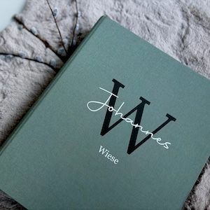 Könnte beinhalten: Ein teal-farbenes Buch mit dem Namen "Johannes" in Schreibschrift und einem großen schwarzen "W" auf dem Einband. Das Wort "Wiese" ist darunter gedruckt. Das Buch liegt auf einer grauen, strukturierten Oberfläche.