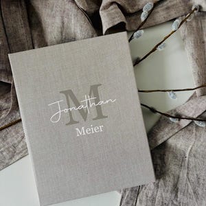 Könnte beinhalten: Ein hellgraues Buch mit dem Namen "Jonathan Meier" auf dem Einband. Der Name ist in einer Mischung aus Schreibschrift und Blockbuchstaben gehalten. Das Buch liegt auf einer weißen Oberfläche mit grauem Stoffhintergrund und einem Zweig mit Kätzchen.