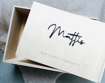 May include: A wooden box with a lid, the lid is open. The box is light brown and has the name "Mattis" written in black cursive on the lid. The text "Meine schönsten Erinnerungen" is written in smaller font below the name.