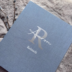 Könnte beinhalten: Ein rechteckiges, mit blauem Stoff bezogenes Album mit einem großen silbernen Buchstaben R, über den der Name Aaron in weißer Schreibschrift geschrieben steht, sowie dem Namen Reinsch in weißen Druckbuchstaben unter dem R.