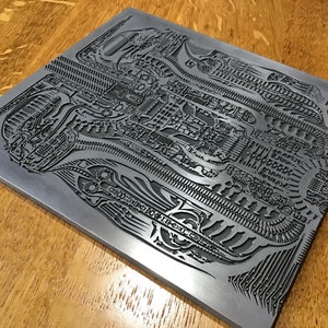 May include: A silver metal plate with a detailed, intricate design etched into the surface. The design features a repeating pattern of lines and shapes, creating a complex and visually interesting texture.