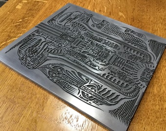 May include: A silver metal plate with a detailed, intricate design etched into the surface. The design features a repeating pattern of lines and shapes, creating a complex and visually interesting texture.