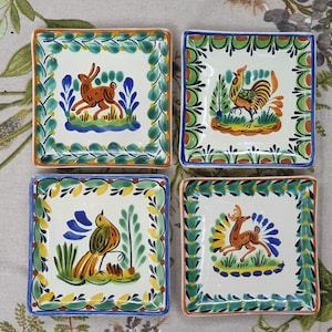 Puede incluir: Juego de cuatro platos cuadrados de cerámica con diseños de animales pintados a mano. Cada plato presenta un animal diferente: un conejo, un gallo, un pájaro y un ciervo. Los platos tienen un fondo blanco con bordes coloridos e ilustraciones de animales.