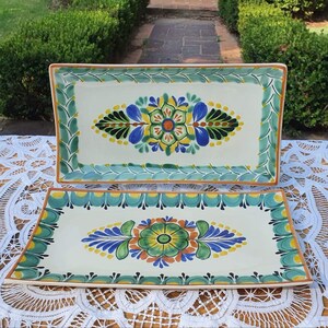 Puede incluir: Dos bandejas de servir de cerámica blanca con diseños florales verdes, azules y amarillos. Las bandejas están sobre un mantel de encaje blanco.