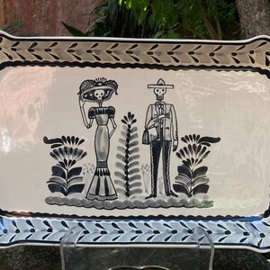 May include: A white ceramic serving tray with a black and gray border and a design of two skeletons in traditional Mexican clothing. The skeletons are standing in front of a floral design.