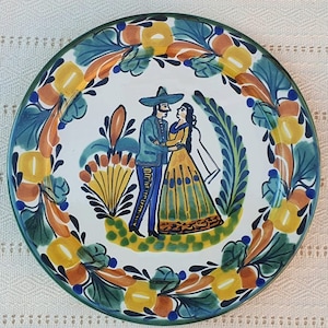 May include: A hand-painted ceramic plate with a traditional Mexican design. The plate features a central image of a couple in festive attire, surrounded by a floral border in shades of blue, yellow, and orange. The plate has a green rim.