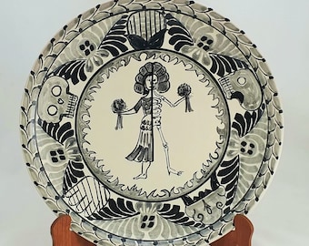 May include: A decorative ceramic plate with a Day of the Dead theme. The plate features a central figure that is half human and half skeleton, holding bouquets. The border has floral and skull motifs in black and gray.