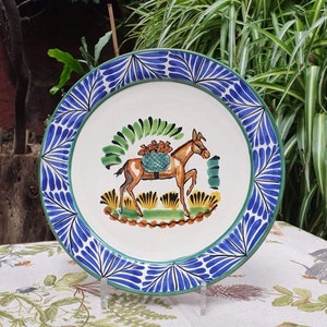 May include: A white ceramic plate with a blue and green border. The center of the plate features a hand-painted illustration of a donkey carrying a basket of fruit. The donkey is brown with a green saddle and a blue basket.