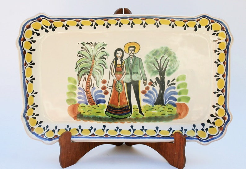 May include: A ceramic serving tray with a hand-painted design of a couple in traditional clothing standing in front of palm trees and a bush. The tray has a yellow, blue, and black border.