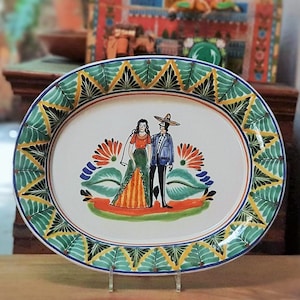 Puede incluir: Plato de servir ovalado de cerámica con un borde verde, amarillo y azul. El centro presenta una escena pintada de una pareja con ropa tradicional mexicana.