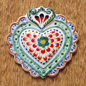 May include: A white ceramic heart ornament with a green, blue, and red floral design. The heart is decorated with a scalloped edge and a small loop at the top for hanging.