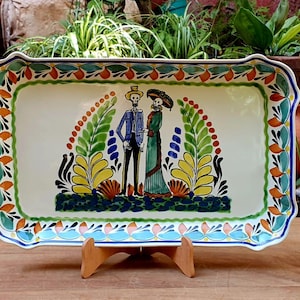 Puede incluir: Bandeja rectangular de cerámica con un colorido diseño del Día de Muertos. La bandeja presenta una base de color crema con una imagen pintada de una pareja de esqueletos, rodeada de motivos florales y de hojas. El borde está decorado con detalles azules, verdes, naranjas y amarillos.