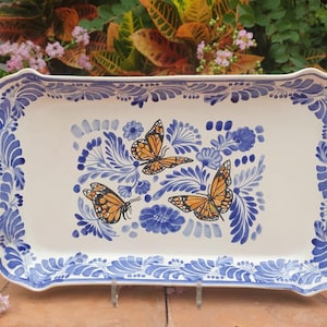 May include: Rectangular ceramic serving tray with a white base and blue floral border. The tray features three orange and black monarch butterflies amidst blue floral designs. The edges have a scalloped design.