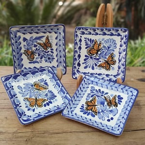 May include: Set of four square ceramic dishes with a white background, blue floral patterns, and orange butterfly illustrations. Each dish has a blue border and is displayed on a wooden surface.