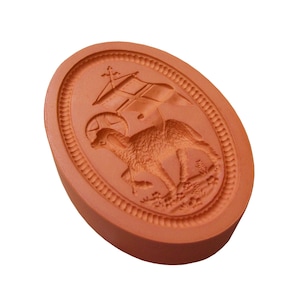 Könnte beinhalten: Ein ovaler Keksausstecher aus Terrakotta mit einem erhabenen Design eines Lammes, das auf einem Sockel mit einem Kreuz und einem Banner darüber steht.