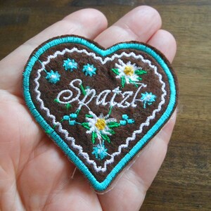 Könnte beinhalten: Ein herzförmiger Aufnäher aus braunem Filz mit dem Wort "Spatzl" in weißer Schrift gestickt. Der Aufnäher hat einen türkisfarbenen Rand und Blumenstickereien in Weiß, Gelb und Blau. Der Aufnäher wird in der Hand einer Person gehalten.