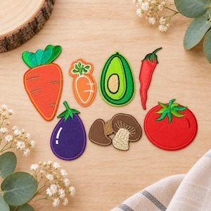 Könnte beinhalten: Eine Sammlung von gestickten Stoffaufnähern in Form von Gemüse und Pilzen, darunter eine große orangefarbene Karotte, eine kleine orangefarbene Karotte, eine grüne Avocado, eine rote Chilischote, eine violette Aubergine, braune Pilze und eine rote Tomate.
