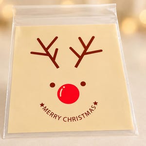 Könnte beinhalten: Eine quadratische cremefarbene Karte in einer transparenten Kunststoffhülle mit einer braunen Rentiergeweih-Grafik, zwei kleinen braunen Augen, einer leuchtend roten runden Nase und dem Text Merry Christmas mit zwei kleinen Sternen.