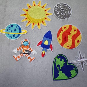 Könnte beinhalten: Ein Set mit neun gestickten Bügelbildern mit Weltraummotiven. Die Patches beinhalten eine Sonne, einen Mond, einen Planeten, eine Rakete, einen Astronauten, eine herzförmige Erde und einen Stern.
