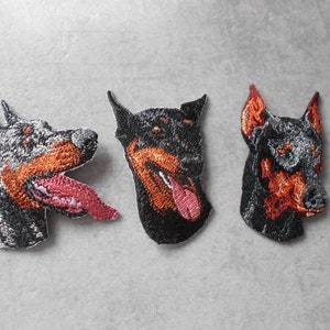 Könnte beinhalten: Drei gestickte Patches von Dobermann-Hunde-Köpfen. Die Patches sind schwarz, braun und grau mit orangefarbenen Akzenten. Die Hunde schauen alle nach rechts.