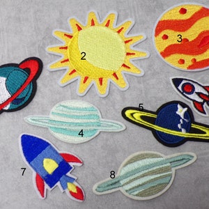 Könnte beinhalten: Acht gestickte Bügelbilder mit einem Sonnensystem, Planeten und Raketen. Die Patches sind von 1 bis 8 nummeriert. Die Sonne ist gelb mit roten Strahlen. Die Planeten sind blau, grün und blau mit gelben Ringen. Die Raketen sind rot und blau.