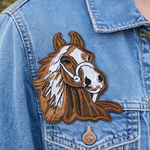 Könnte beinhalten: Ein brauner und weißer Pferdekopf-Aufnäher ist auf eine hellblaue Jeansjacke genäht. Der Aufnäher zeigt detaillierte Stickereien des Gesichts des Pferdes, einschließlich eines Zaumzeugs. Die Jacke hat eine Knopfleiste und eine Brusttasche.