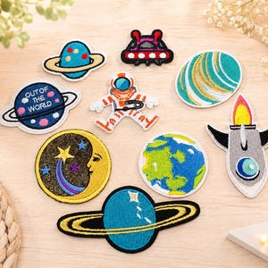 Könnte beinhalten: Eine Sammlung von neun bestickten Aufnähern zum Thema Weltraum, darunter ein blauer Planet mit Ringen, ein dunkelblauer Planet mit dem Text OUT OF THE WORLD, ein rotes und schwarzes UFO, ein Astronaut, ein gestreifter Planet, eine Mondsichel mit Gesicht, die Erde, eine Rakete und ein blauer Planet mit goldenen Ringen.