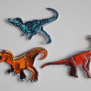 Könnte beinhalten: Drei gestickte Bügelbilder mit Dinosaurier-Motiven. Die Patches sind in den Farben Blau, Orange und Rot gehalten.