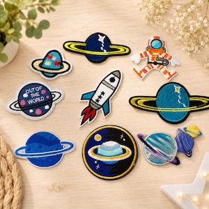 Könnte beinhalten: Eine Sammlung von neun bestickten Aufnähern zum Thema Weltraum mit verschiedenen Planeten, einer Rakete und einem Astronauten. Die Designs umfassen einen blauen Planeten mit dem Text "OUT OF THE WORLD", eine weiße Rakete und Ringplaneten.