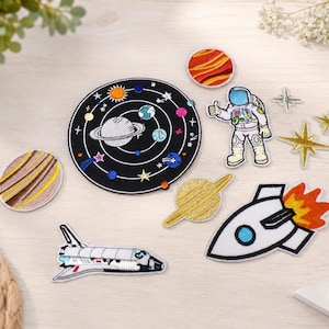 Könnte beinhalten: Eine Sammlung bestickter Aufnäher zum Thema Weltraum, darunter eine große kreisförmige Karte des Sonnensystems, ein Astronaut, ein Space Shuttle, eine Rakete mit Flammen sowie verschiedene Planeten und Sterne in leuchtenden Farben.
