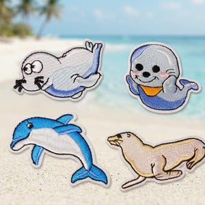 Könnte beinhalten: Vier gestickte Aufnäher mit Meerestieren. Enthält einen Cartoon-Seehund mit großen Augen, einen lächelnden Seehund mit Lätzchen, einen blau-weißen Delfin und einen beigefarbenen Seehund. Alle Aufnäher haben einen weißen Rand.