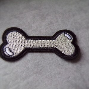 Könnte beinhalten: Gestickter Knochen-förmiger Aufnäher mit schwarzem Rand und hellgrauer Füllung. Der Aufnäher hat ein detailliertes, strukturiertes Aussehen mit weißen Akzenten an den Enden. Geeignet für Bastelarbeiten und Kleidung zum Thema Haustiere.