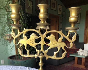 Può includere: Un candelabro in metallo dorato con tre portacandele e una base decorata con figure di leoni stilizzate. Il candelabro ha un design tradizionale ed è adatto a una varietà di stili di arredamento per la casa.
