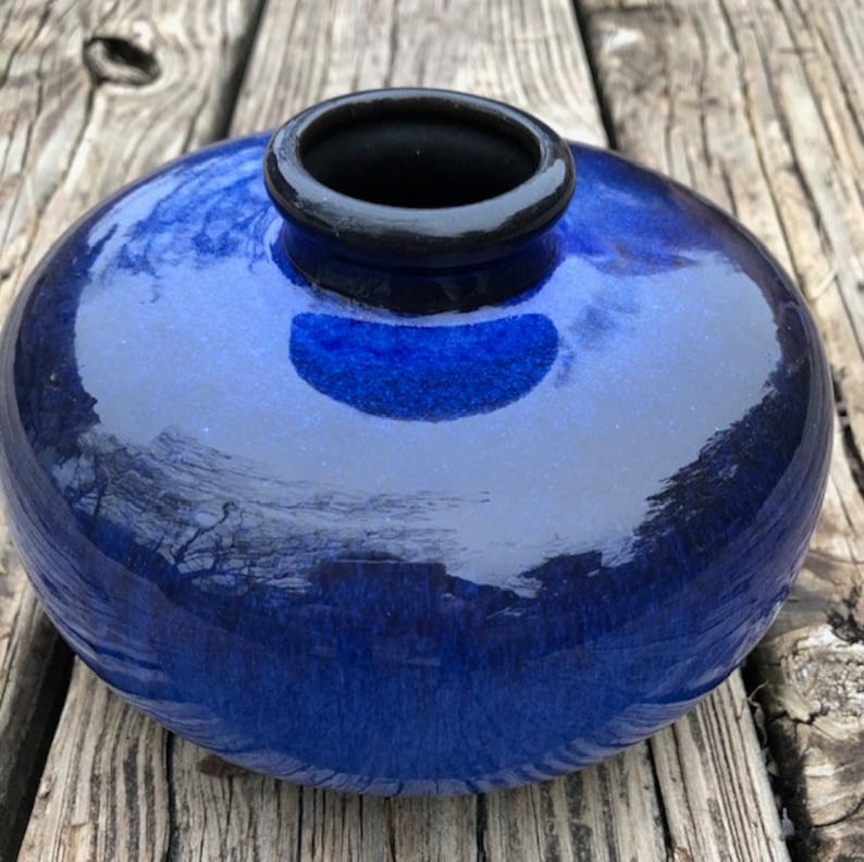 May include: A small, round, blue ceramic vase with a black rim. The vase has a glossy finish and is perfect for displaying small flowers or greenery.