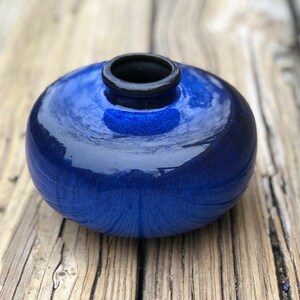 May include: A small, round, blue ceramic vase with a black rim. The vase has a glossy finish and is sitting on a wooden surface.